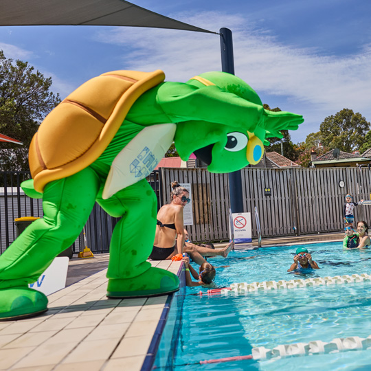 Aquatics mascot Terry the Turtle posing on the edge of the outdoor pool at FDAC as though he is about to dive into the water